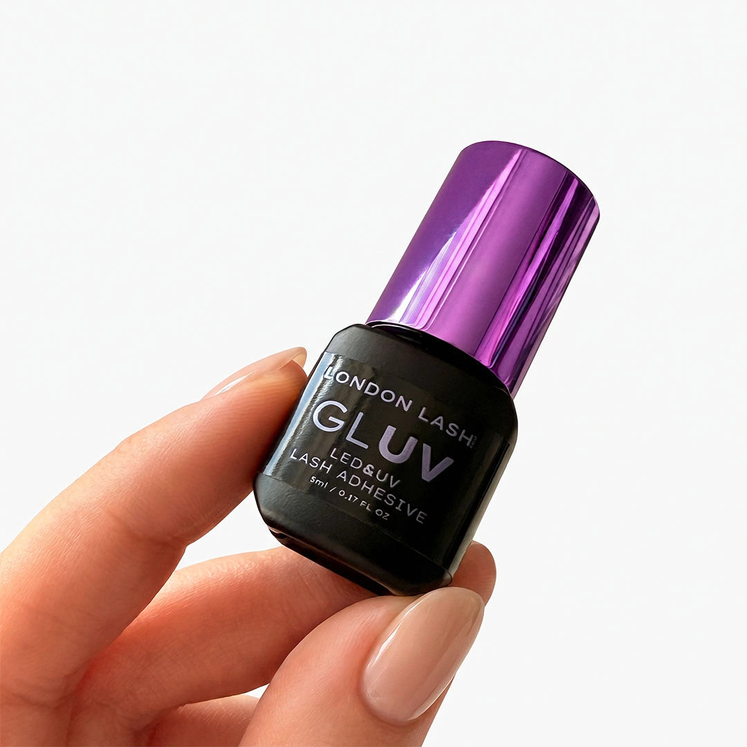 Gros plan sur une main manucurée tenant le flacon de 5 ml de la colle London Lash GLUV, doté d'un bouchon violet brillant.