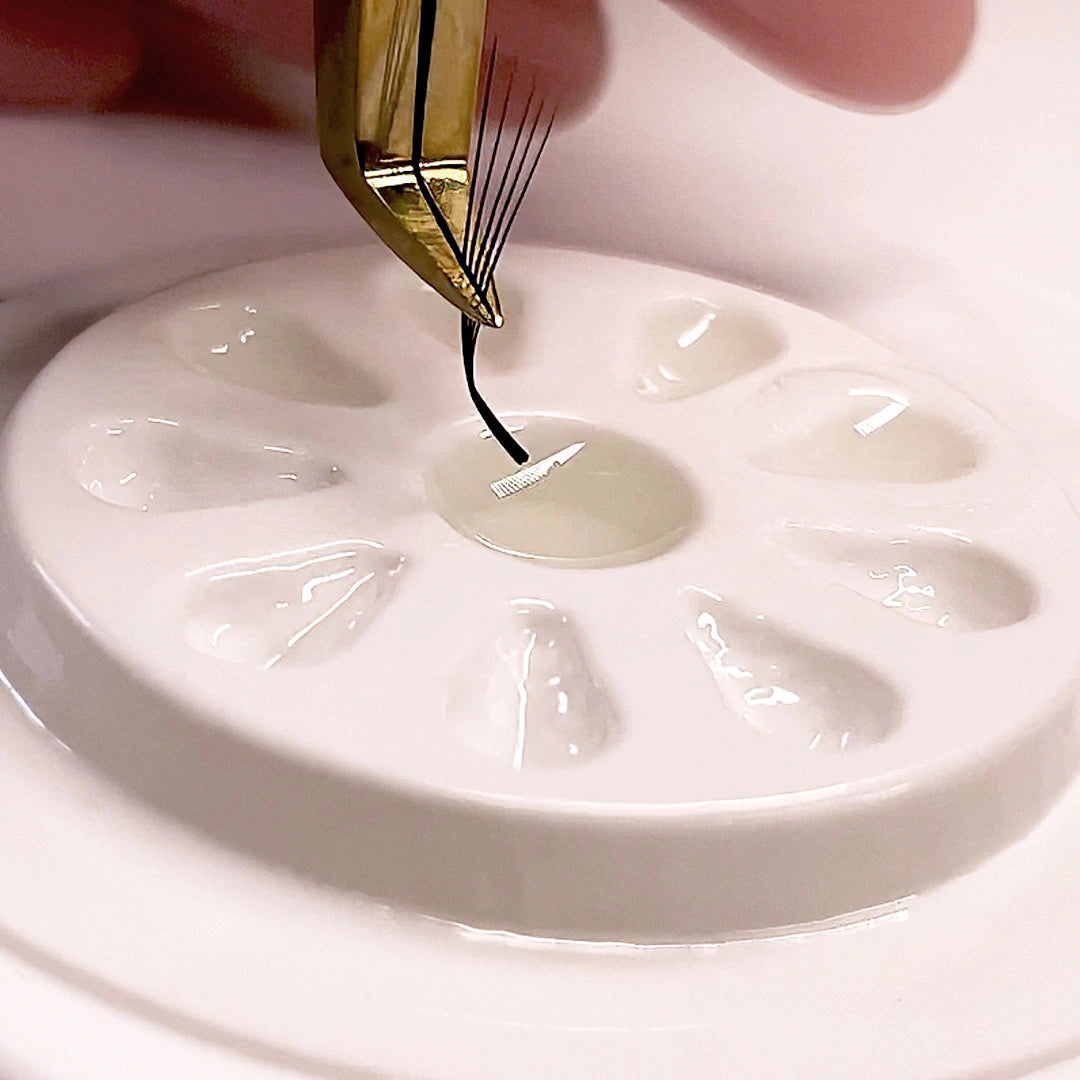 Photo macro d'une pince dorée trempant la base d'un éventail de volume pour extensions de cils dans de la colle transparente sur une palette de colle blanche.