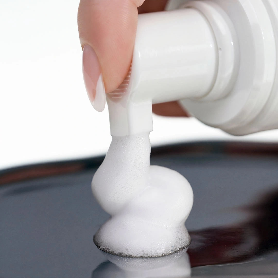 Gros plan de la pompe distribuant une mousse nettoyante blanche et onctueuse sur une surface métallique réfléchissante.